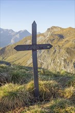 A rusty, iron cross on a mountain peak with an impressive mountain landscape and clear sky in the