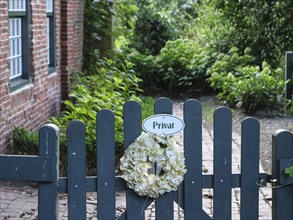 A garden fence with a 'Private' sign, decorated with a wreath of flowers, surrounded by plants,