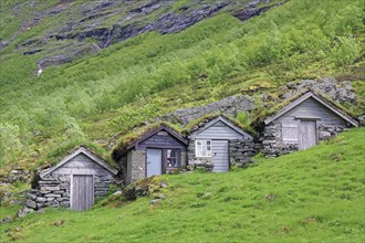 Three small rustic wooden cabins nestled in a green hilly landscape with grass and rocky mountains