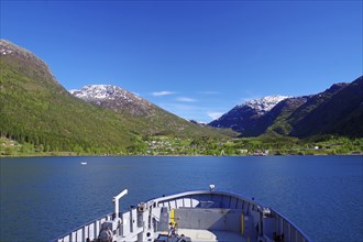 View from a ferry onto a Norwegian fjord surrounded by snow-capped mountains and blue sky,