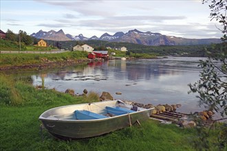 An abandoned boat lies on a grassy shore with mountains and houses in the background, Saltstraumen,