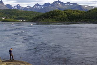 A man stands on the bank of a tidal stream, surrounded by green hills and mountains in the