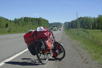 A bicycle loaded with panniers stands on the side of the road in a green, rural landscape under a