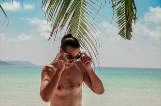 A shirtless man with a hair bun adjusts his sunglasses under palm leaves on a tropical beach with