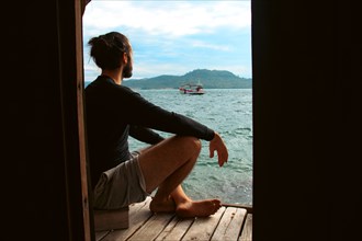 A man sitting on a dock looking out at the sea and a boat with mountains in the background