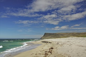 A sunny beach with deep blue water and waves, surrounded by cliffs and bright skies, Latrabjarg,