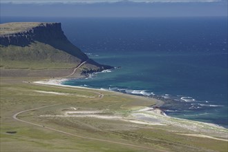 A cliff juts out over the deep blue sea, next to a long, sandy beach and green meadows, Latrabjarg,