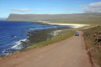 A car drives along a coastal road passing a rugged coastline with blue water, Latrabjarg,