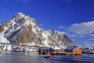 Harbour with bridge, surrounded by snowy mountains, boats in the water and sheds on the shore,