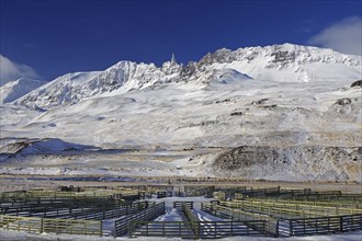 Wooden fences in a circular arrangement in front of a snow-covered mountain range under a blue sky,