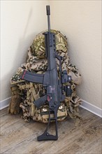 Rifle and military equipment with camouflage pattern, including a rucksack, placed in front of a