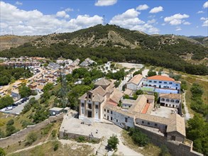 Image of a town with a church in front of a wooded hill under a blue sky with clouds, Ermita de San