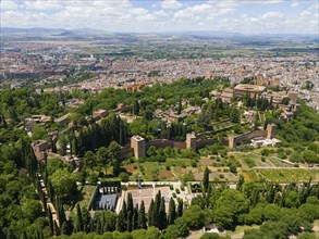 Aerial view of a castle complex, embedded in green surroundings and surrounded by a city and hills,