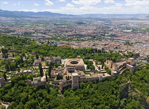 Aerial view of a castle complex surrounded by green trees and hills with a city in the background,