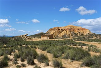 Small hut in front of a large rocky hill in a desert-like landscape under a blue sky, Bardenas