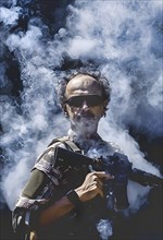 A mercenary in uniform and sunglasses stands in the powder smoke with his assault rifle, AI