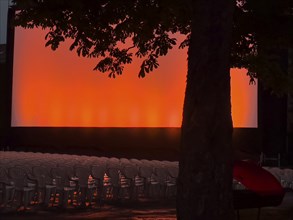 Rows of chairs and a tree in front of a red and orange illuminated screen in the open-air cinema