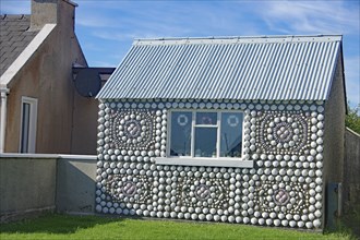 House facade decorated with shells, including a window, next to another building and garden,