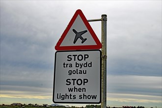 Road sign with an aeroplane symbol and bilingual text in a rural landscape under a cloudy sky,