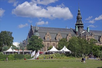 Historic building with striking architecture next to a green space and people under a clear sky,