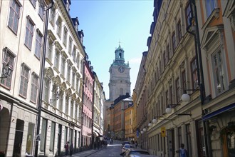 Narrow street between tall, pastel-coloured buildings with a view of a church tower on a sunny day,