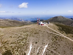 Hilly landscape with radar towers, rugged paths and a wide view under a slightly cloudy sky, aerial