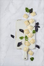 Overhead view of raw uncooked ravioli on a chopping board with scattered leaves of basil and