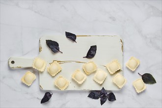 Overhead view of raw uncooked ravioli on a chopping board with scattered leaves of basil