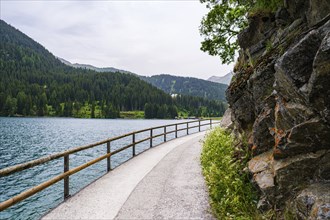 A winding path along the lake with a wooden railing and rocky side, surrounded by wooded mountains,