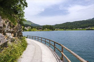 Path with railing along a lake, surrounded by mountains and forests under a slightly cloudy sky,