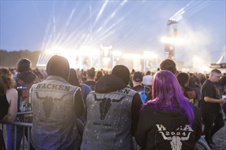 Festival visitors with Wacken jackets look at the two main stages Faster and Harder at dusk at the
