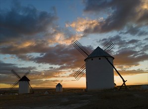 Three windmills in front of a dramatic sunset with a colourful sky and clouds. The silhouettes of