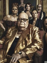 Elderly man in a gold jacket seated in an ornate room with serious individuals standing behind,