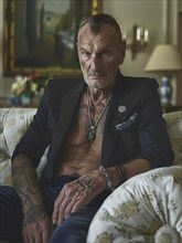 Elderly man with tattoos in a blazer sitting on a rustic sofa, with an intense, serious gaze, AI