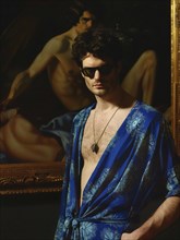 Man in a blue robe and sunglasses standing in front of an artistic painting, exuding a mysterious