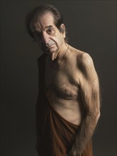 An elderly man, shirtless and draped in brown cloth, stands against a dark background with a solemn