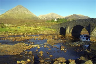 Historic stone bridge over a river with mountains in the background, surrounded by green meadows