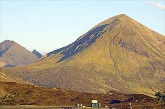 Large single mountain with rocky surface illuminated by yellow sunlight under a clear blue sky,