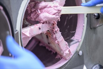 Workers in gloves scrape pink ice cream from a machine in an industrial factory setting,