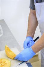 Person cutting a cantaloupe in the kitchen while wearing gloves, showing food preparation and hand