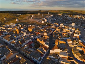 Roofs and houses of a village at dusk with windmills in the background and orange lighting, aerial