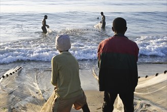 Boys are pulling a fishing net out of the water. Two fishermen are helping them. At Toamasina