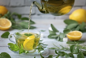 Tea is poured from a glass jug into a glass cup, surrounded by lemons and herbs