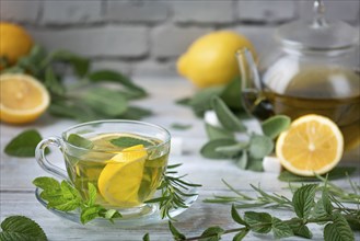 Herbal tea in a glass cup and glass jug surrounded by herbs and lemon slices