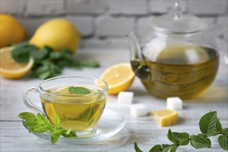 A glass cup with tea, mint and lemon, next to sugar cubes and a glass jug on a wooden table