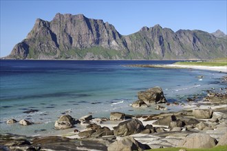 Wide beach with clear blue water and rocks, while impressive mountains form the background,