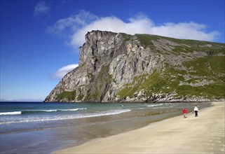 Two people walking along a wide sandy beach with high cliffs and clear water, Kvalvika, Fredvang,