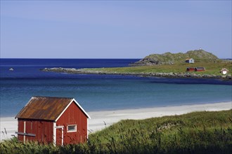 Red wooden house on a sandy beach with a view of the clear blue water and green island, Ramberg,