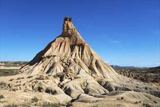 An imposing rock formation in the middle of a desert-like area under a light blue sky,