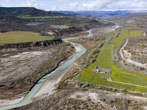 Aerial view of a river winding through a green meadow and past ancient ruins in the valley,